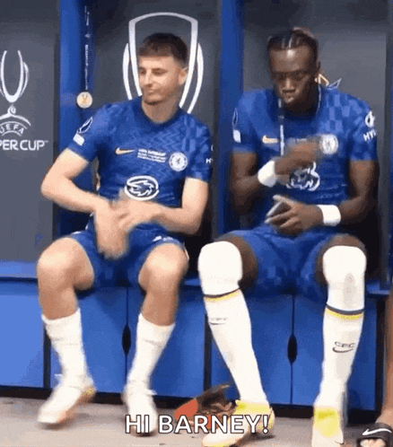 two soccer players are sitting on a bench in a locker room and one of them is talking to the other .
