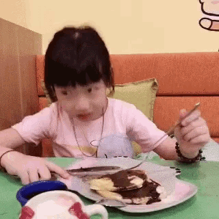 a little girl is sitting at a table with a plate of food and a fork .