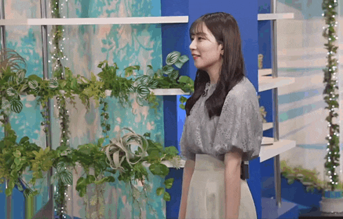 a woman in a grey top and white skirt stands in front of a blue wall surrounded by plants