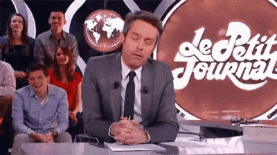 a man in a suit and tie is sitting at a desk in front of a sign that says le petit journal