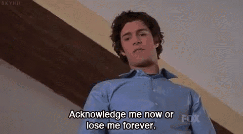 a man in a blue shirt is standing in a room and saying `` acknowledge me now or lose me forever '' .