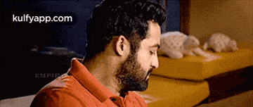 a man with a beard wearing a red shirt is sitting on a couch looking at something .