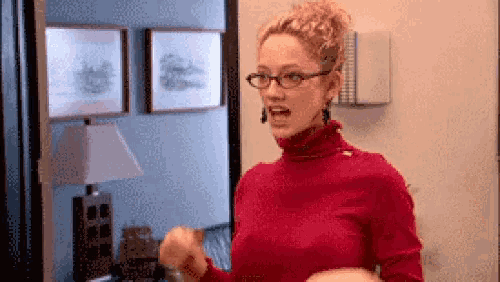a woman wearing glasses and a red turtleneck is standing in a room with a lamp .