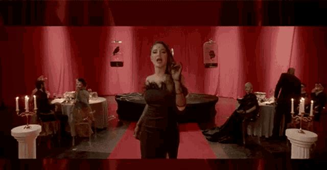 a woman in a black dress is standing on a red carpet in a room with people sitting at tables and candles .