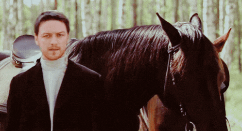 a man in a suit stands next to a horse in the woods