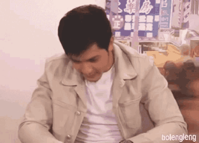 a man in a tan jacket and white shirt is sitting at a table with his head down .