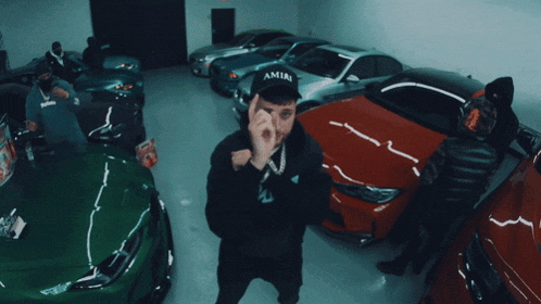 a man wearing a black amiri hat stands in front of many cars