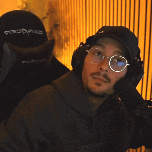 a man wearing glasses and headphones holds his finger to his mouth in front of a dxracer chair