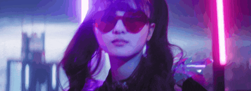 a woman wearing sunglasses and a purple jacket is standing in front of a neon light .
