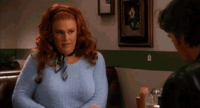 a woman with red hair wearing a blue sweater and a scarf talks to a man