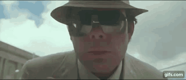 the man is wearing a hat and sunglasses and looking at the camera .