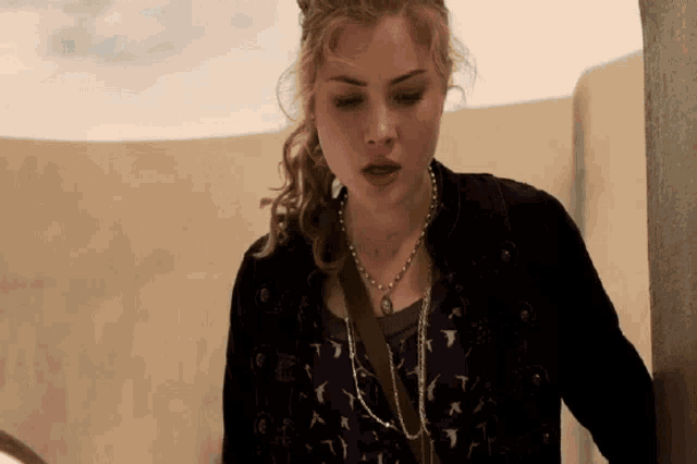 a woman wearing a black jacket and a necklace is standing in front of a wall