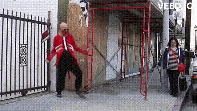 a man in a red jacket is dancing on a sidewalk in front of a fence and scaffolding ..