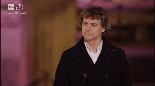 a man in a black coat stands in front of a pink background that says rai on it