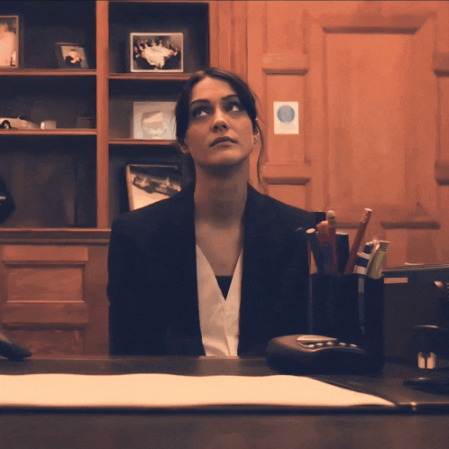 a woman in a suit sits at a desk in front of a wall with pictures on it
