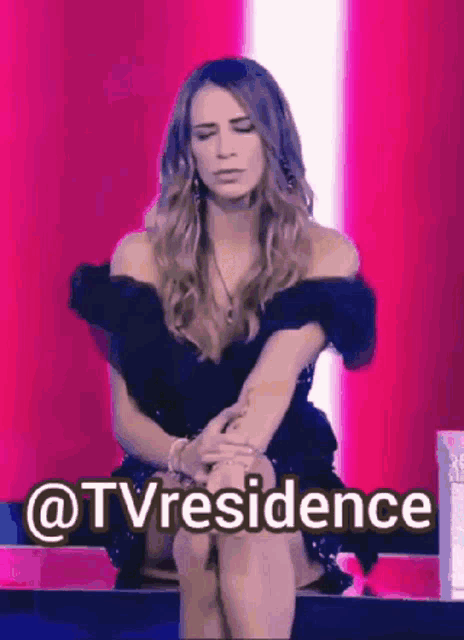 a woman in a black dress is sitting on a bench with her knees crossed and a pink background .