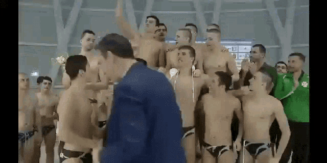 a man in a suit is standing in front of a group of naked men in swimming suits .