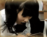 a woman with long black hair is sitting at a table with a phone in her mouth .