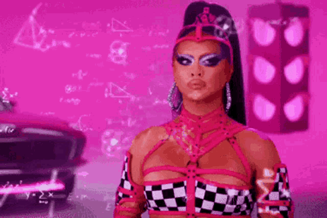 a drag queen in a pink and black checkered outfit is standing in front of a pink wall .