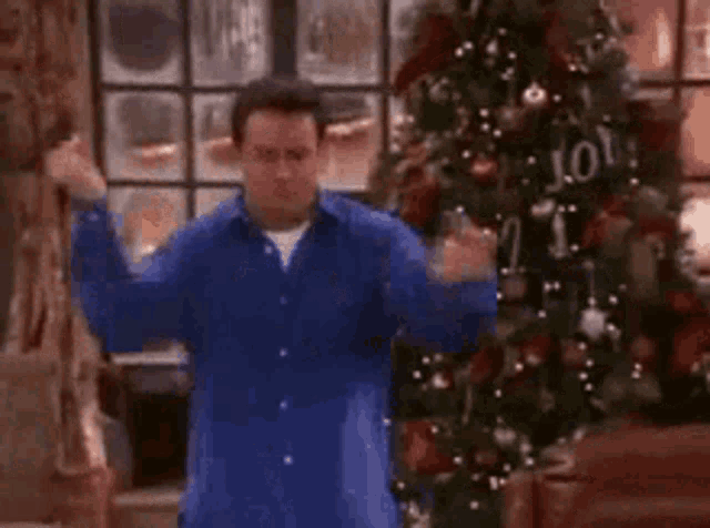 a man in a blue shirt is dancing in front of a christmas tree in a living room .