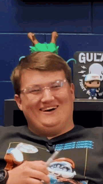 a man wearing glasses and a black shirt has a green toy on his head in front of a sign that says gula