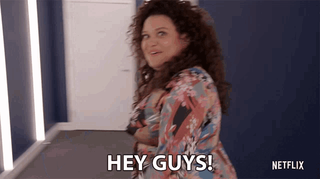 a woman with curly hair says hey guys in a hallway
