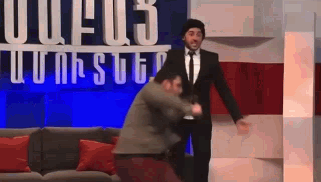 a man in a suit and tie is standing next to another man in a couch .