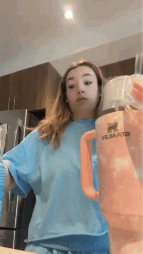 a woman in a blue shirt is standing next to an orange stanley tumbler
