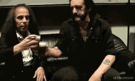 two men are sitting next to each other on a couch toasting with beer .