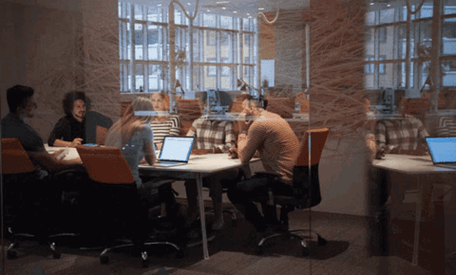 a group of people are sitting around a table with laptops in front of a glass wall