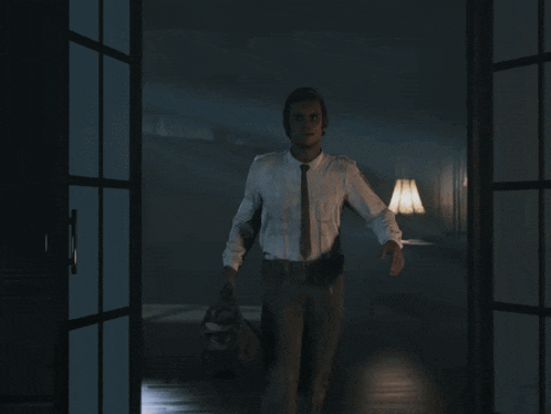 a man in a white shirt and tie is standing in a dark room with a lamp behind him