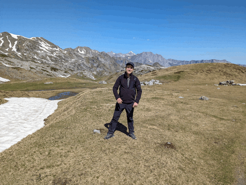 a man in a black jacket stands on a grassy hill with mountains in the background