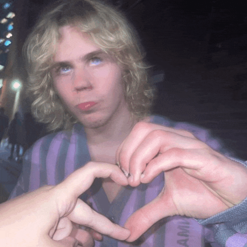 a person making a heart shape with their fingers in front of a man with blonde hair