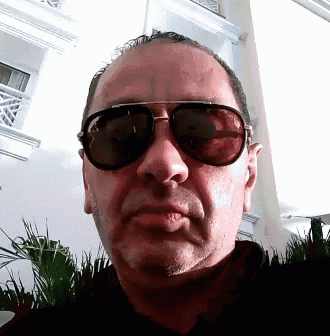 a man wearing sunglasses looks at the camera with a serious look on his face