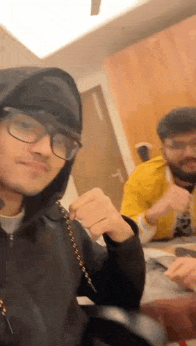 a man wearing glasses and a hoodie is taking a selfie with another man while sitting at a table .