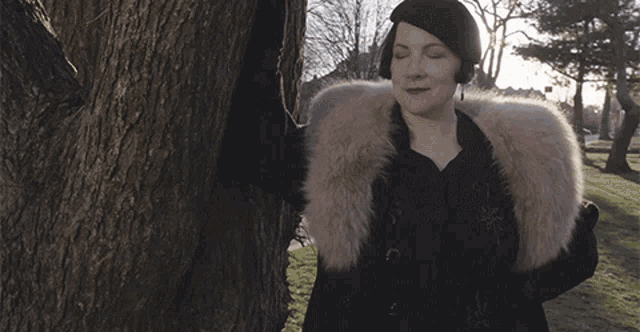 a woman leaning against a tree wearing a fur coat