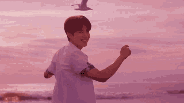 a young man is standing on a beach at sunset with a seagull flying in the background .