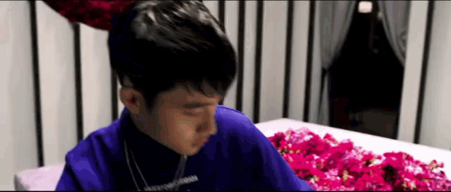 a man in a blue robe is sitting in front of a pink coffin covered in flowers .