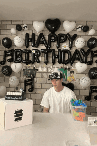 a man stands in front of a wall with balloons and a cake that says happy birthday