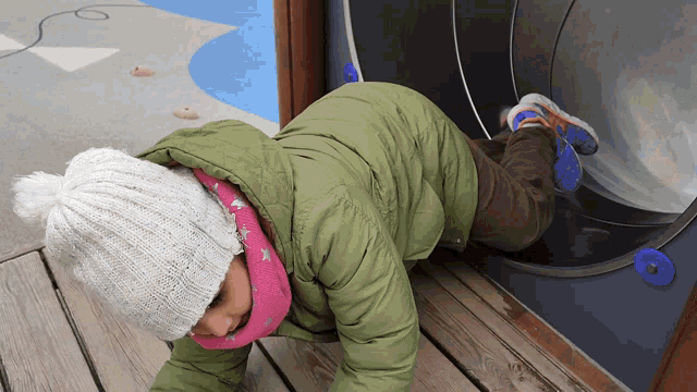 a child wearing a green jacket and a white hat is crawling on a wooden deck