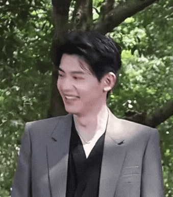 a young man in a suit and black shirt is smiling in front of trees .