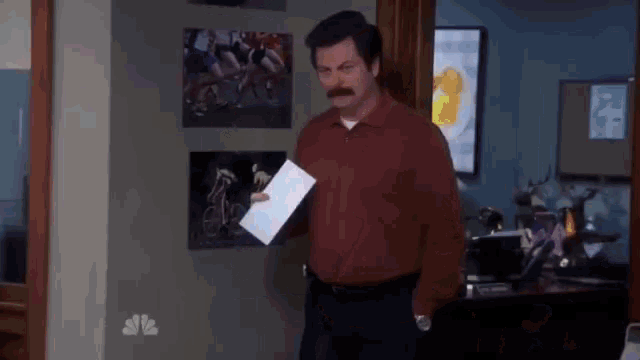 a man in a red shirt is holding a piece of paper in his hand in a room .