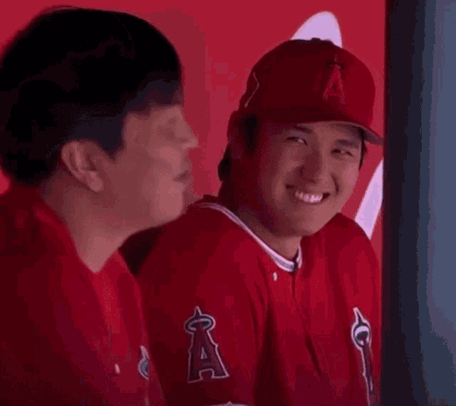 two baseball players are sitting next to each other in the dugout and smiling .