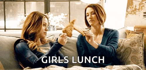 two women are sitting on a couch eating food and the words girls lunch are on the bottom .