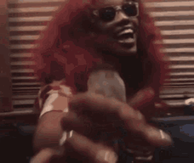 a woman with red hair and sunglasses is sitting on a couch holding a bottle of wine .