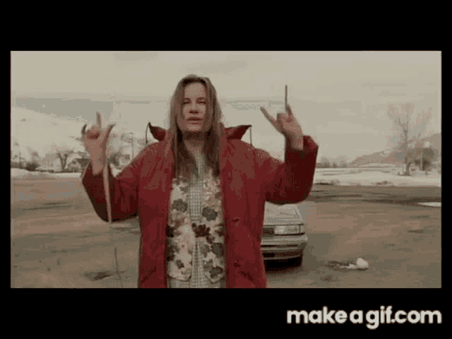 a woman in a red jacket is standing in a parking lot with her arms outstretched and making a peace sign .
