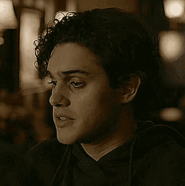 a young man with curly hair is wearing a black hoodie and looking at the camera in a dark room .