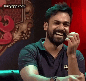 a man with a beard is sitting on a couch laughing with his hands in his mouth .