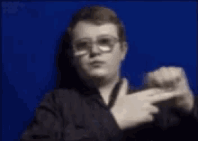 a man wearing glasses is pointing at something in front of a blue wall .