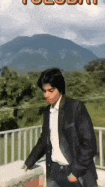 a man in a suit and white shirt is standing on a balcony with mountains in the background .